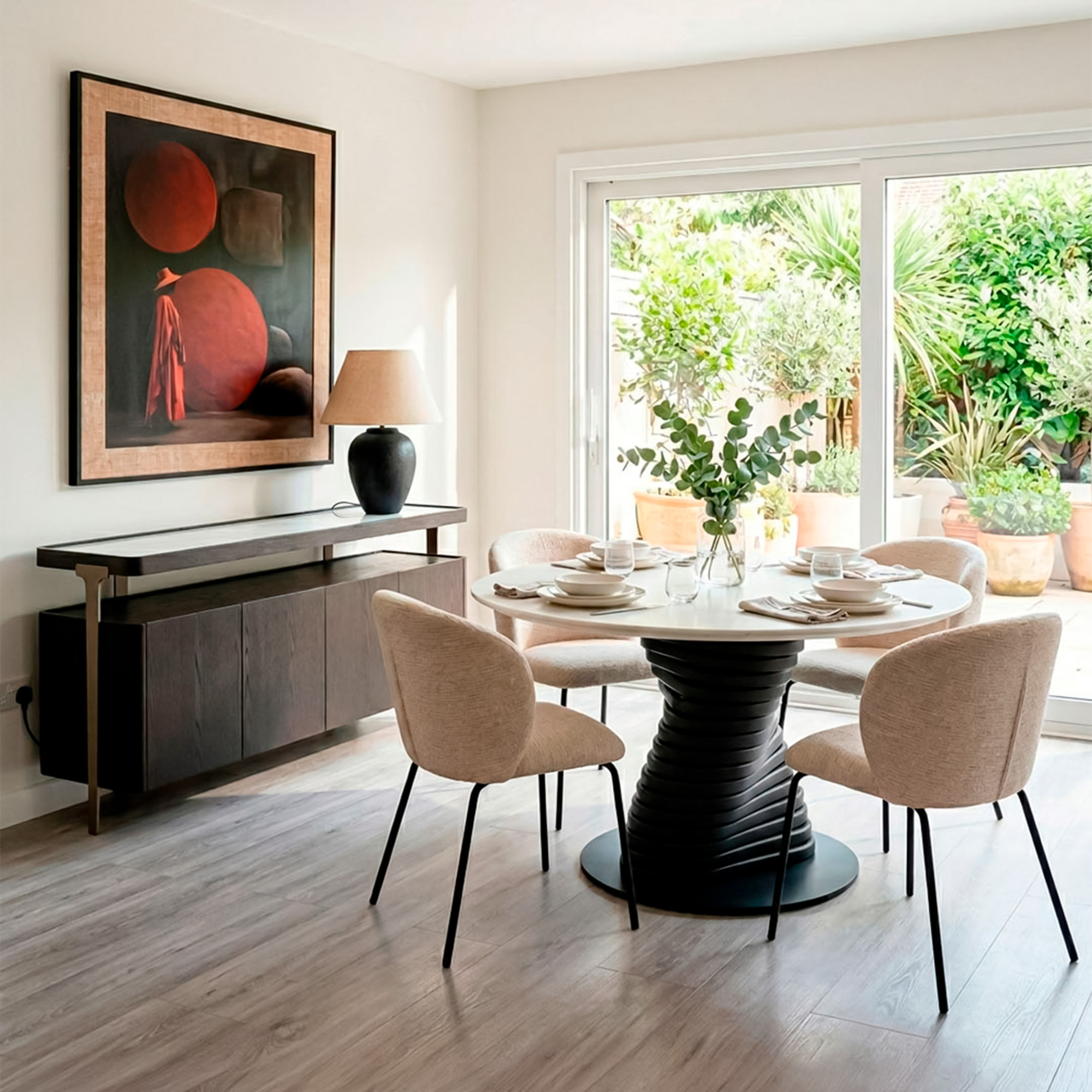 Bright dining room with a round table featuring a ribbed black pedestal base and light stone top, four beige fabric chairs with black legs, dark wood sideboard with metal details and black ceramic table lamp